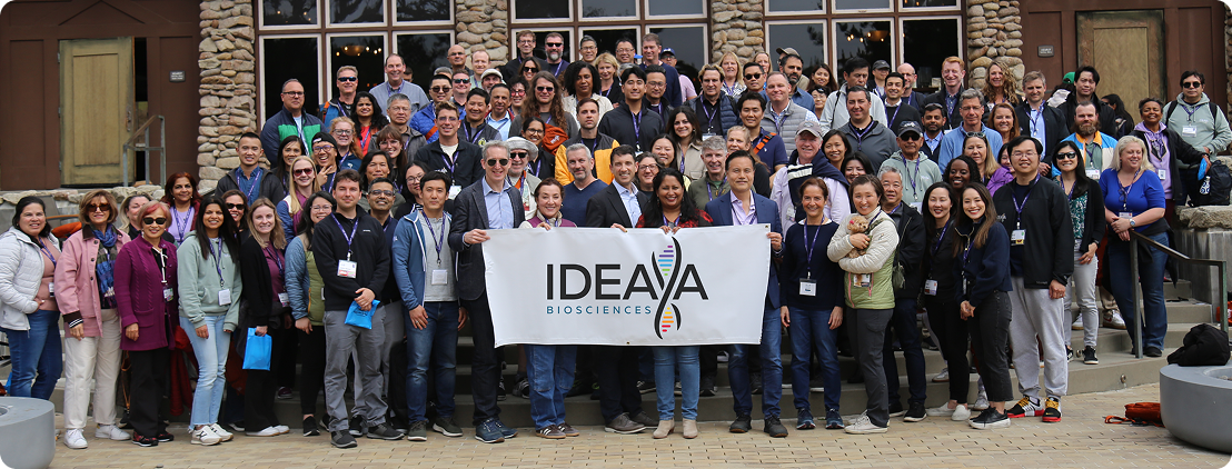 Large group of people smiling outside, holding a banner that reads "IDEA" in front of a building.