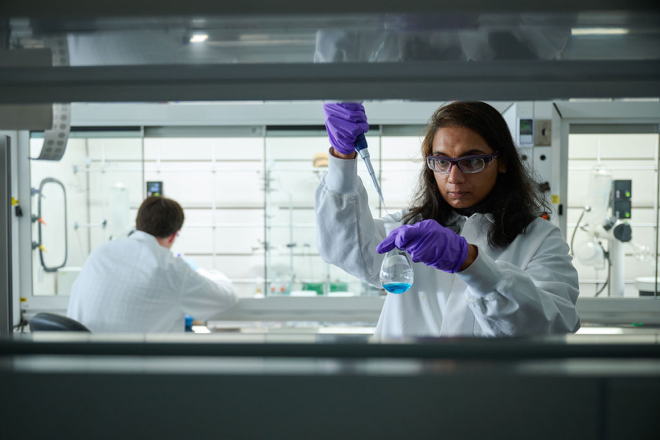 Scientist in lab gloves examines liquid in beaker, another works in background.